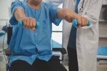 Asian older aged man doing physiotherapist with support from nurse. Senior elderly male sitting on sofa in living room using dumbbells workout exercise for patient with caregiver in nursing care.