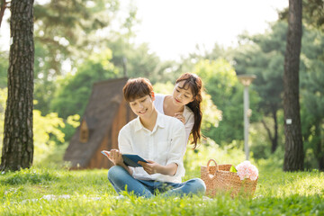 A young male and female couple are enjoying a pleasant picnic while drinking drinks and reading books on the forested park lawn.