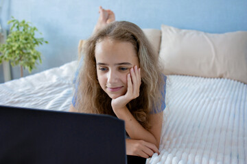 Fototapeta premium A teenage girl watches videos on a laptop or reads student chats while lying on her bed in the bedroom.