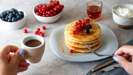 Pile of pancakes with red currants, blueberries, honey, cream cheese, cultery and napkin over grey background. Two hands holding cup of coffee and fork