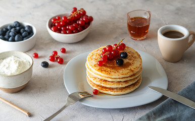 Pile of pancakes with red currants, blueberries, honey, cream cheese, cultery and napkin over grey background