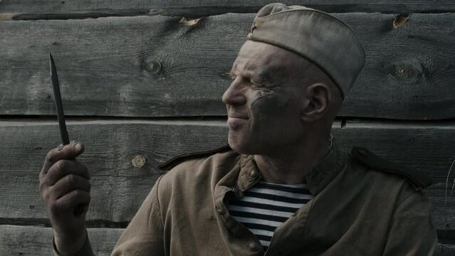 Medium close-up portrait of mature soviet soldier with dust and mud on face sitting in frontline trench checking sharpness of combat knife