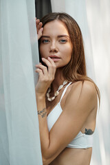 portrait of woman with tattoo and blue eyes posing in bikini top near white tulle of beach house