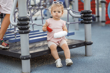 Adorable toddler with a broken arm of a child in a cast sitting on the playground. Girl a 2-3-year-old keeps her arm bent against the background of a pink T-shirt. Trauma after visiting the playground - obrazy, fototapety, plakaty