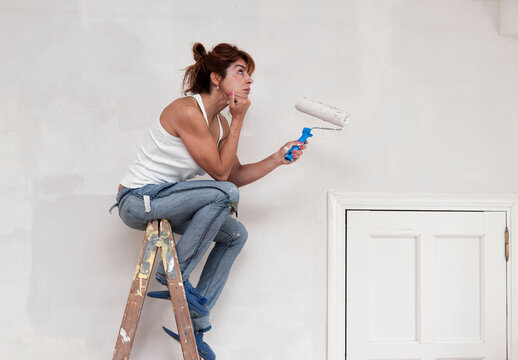 A Woman Sits At The Top Of A Ladder With A Paint Roller In Her Hand; Tarifa, Cadiz, Andalusia, Spain