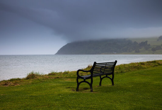 A bench sits on the water's edge with storm clouds over the water; Moray firth scotland