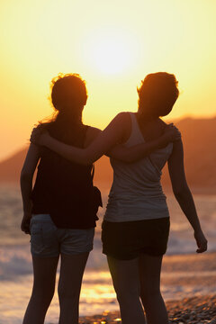 Two friends in an embrace as they watch the sunset at the water's edge; Aphrodite bay, cyprus