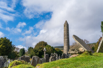 Fototapeta na wymiar Medieval ruined graveyard with round tower in sunshine at churchyard in Glendalough's monastic town. Early medieval monastic settlement founded by St Kevin in the 6th century
