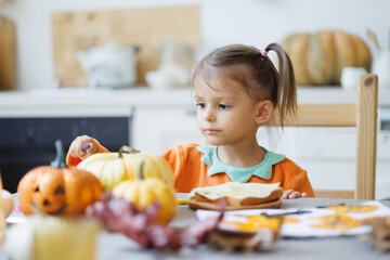Happy Halloween! Little cute girl eating cookies in the kitchen. Toddler girl in a pumpkin costume eating sandwiches on breakfast. Morning at home.