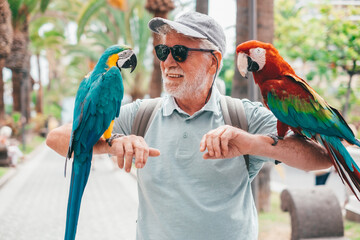 Smiling elderly bearded man in hat and sunglasses holding on his arms two domestic ara parrots in the park © luciano