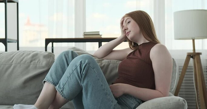 A red haired teenage girl is shown in the living room, appearing bored and disengaged. The subdued lighting and the casual setting of the living room emphasize the feeling of monotony.
