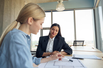 Close up of two female design engineer working with project documents sitting in office near window
