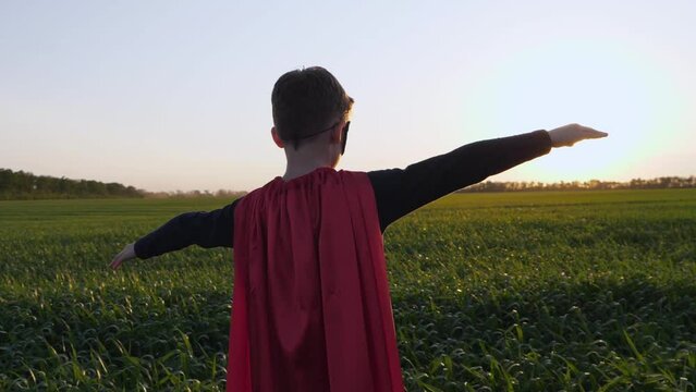 A happy child in a red superhero cape stands against the background of the sky and a green meadow with his arms outstretched to the side depicting flight. The concept of a kid dream.