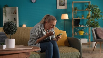 A woman holds a phone and screams with joy. A woman won an online game or received a prize. Portrait of a happy, shocked woman with a phone in her hands on the sofa in the living room.