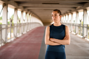 Sporty woman dressed in activewear looking away with thoughtful face expression and arms crossed poses on bridge