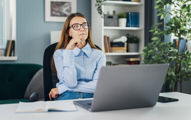 Obraz premium Young thoughtful woman wearing eyeglasses dreaming at desk with laptop, motivated idea concept, working in cozy light room