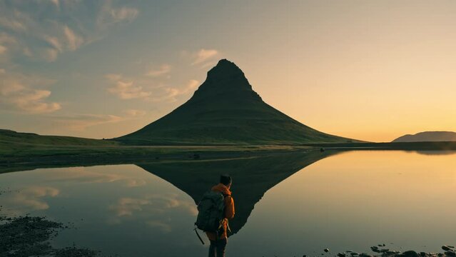 Beautiful landscape of sunrise over volcanic Kirkjufell mountain with lake reflection and male backpacker enjoying on coast in summer at Snaefellsnes peninsula, Iceland