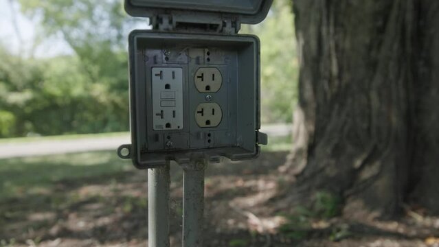 Handheld slow motion of a North American outdoor power outlet with electrical current and plugs sticking out of the ground next to tree in back yard garden and lawn