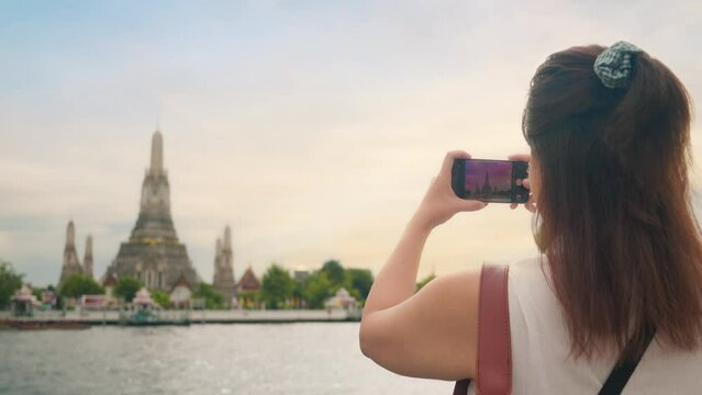 Back view of young Asian woman tourist using smartphone taking photo of Wat Arun Ratchawararam Temple of Chao Phraya River, Bangkok Thailand
