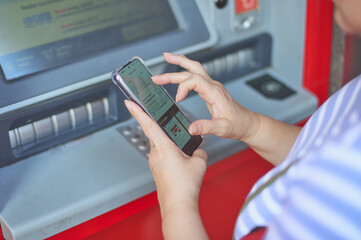 Unrecognizable woman holding a cell phone in front of a bank ATM carrying out an online banking operation