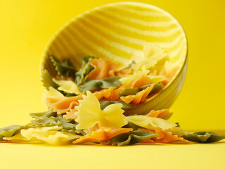 Close up, colorful pasta in the shape of a bow tie on yellow background and lying bowl.