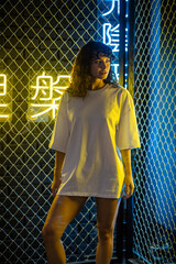 Attractive woman with curly hair in a white oversized t-shirt stands against the background of neon signs.