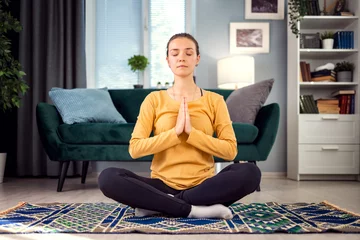 Tuinposter Front view of pretty young woman sitting in lotus pose at home floor, recreation and relax concept © MYDAYcontent