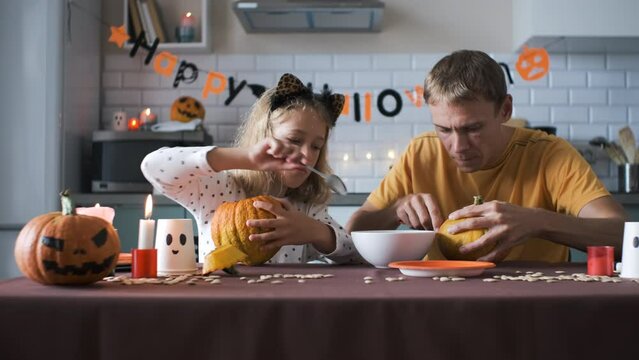 Father with little daughter carving jack-o-lanterns in the kitchen, slow motion. Preparing for Halloween celebration
