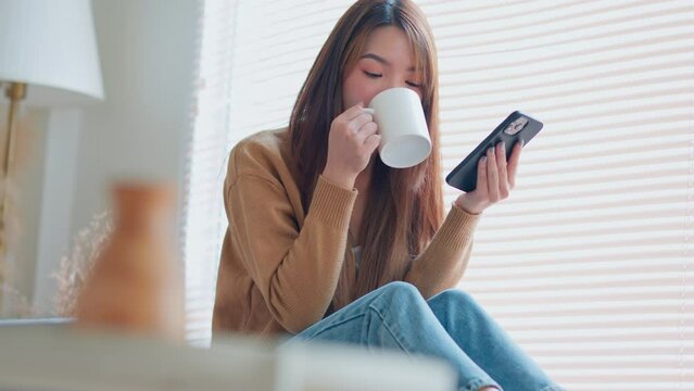 Relaxed young asian woman using mobile smartphone and drinking coffee from a mug while sitting by the window at home, The concept of balance and tranquility.