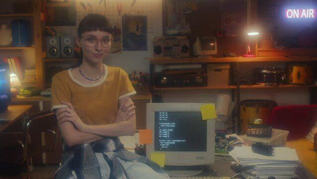 Medium portrait of young female programmer sitting at desk in garage office equipped with old computers crossing her arms looking at camera