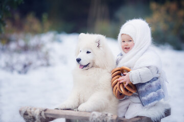 Small girl with bagels sitting on sledges with white dog in winter park