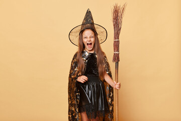 Crazy girl dressed in witch costume holding broom isolated over beige background having fun at...