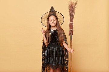 Hopeful girl dressed in witch costume holding broom isolated over beige background posing at halloween party keeps fingers crossed making wish desire. - obrazy, fototapety, plakaty