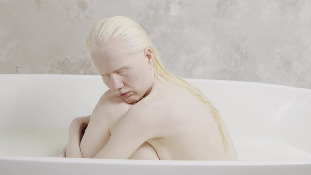 Portrait of melancholic albino woman with wet hair sitting with eyes closed in bathtub hugging her knees