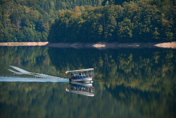 A small boat glides across the smooth surface of lake. The shores with dense forest