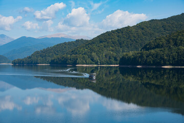 Small boat glides across the surface of lake surrounded by green forest hills and amazing reflection of cloudy sky. Vidrarudam. Trasfagarasan road. Romania