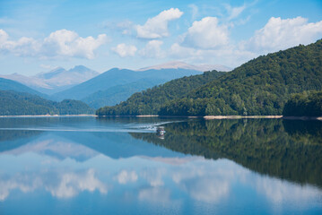 Vidraru lake with dense forest around and a small boat glides across the surface of lake. Beautiful reflection of cloudy sky in the calm weather