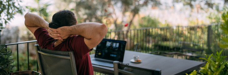 A young, handsome man is sitting relaxed at a laptop at a table on a veranda in a green garden.