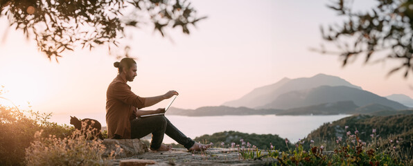 A Caucasian man is working with a laptop in a garden on a mountain overlooking the sea and sunset.