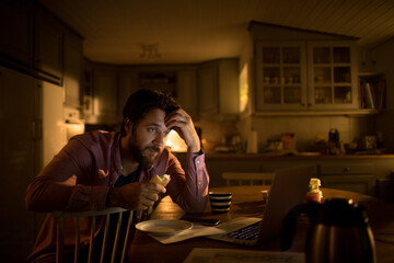 Worried Young Caucasian man having a snack while going over bills and home finances at night at home