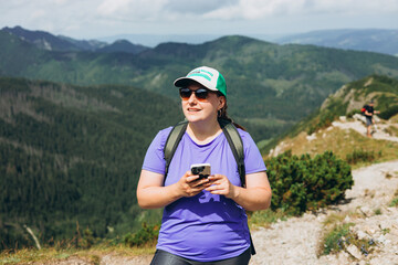 Happy woman hiker with backpack standing on the slope of mountain ridge against mountains. 30s Women holding smart phone. Travel and active lifestyle concept, outdoor activities