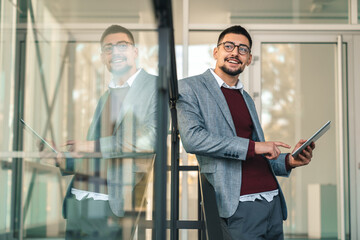 Naklejka na ściany i meble Young happy businessman company manager using digital tablet outside modern work office. Smiling male business professional with eyeglasses wearing smart casual clothes leaning on glass office.