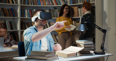 Young Caucasian man in VR headset sitting in library space, scrolling and moving hands in air and having virtual reality experience.