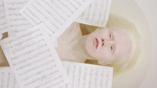 Top-down portrait of young female musician with albinism lying in bathtub with sheet music papers on water covering her body