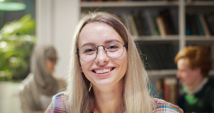 Close up of pretty joyful young blonde Caucasian female student in glasses looking at camera and smiling. Indoor. Beautiful woman studying in library. Portrait shot.