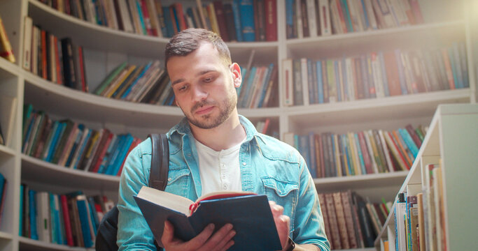 Young handsome Caucasian male student in with backpack standing in library, reading textbook and closing it. Putting book on shelf and leaving. Literature concept. Bookshelves background.