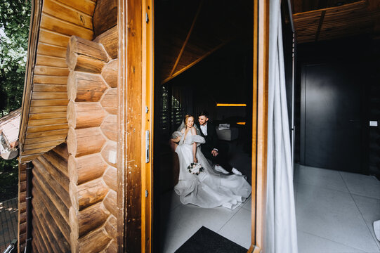 Stylish young groom in a green suit and a beautiful smiling bride in a white dress are hugging while sitting on a sofa, bed in a hotel room. Wedding photography, portrait, lifestyle.