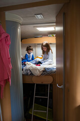 Two caucasian children, brother and sister, reading books in pajamas on a campervan bed during a road trip stop. Camper vacations travelling with kids. vertical shot.