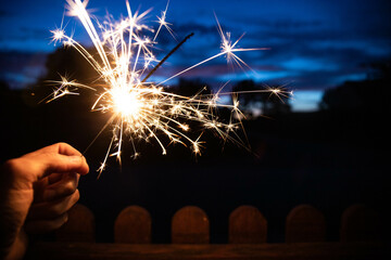 Hand holding a sparkler, or bengal fire stick, burning in outdoor setting at blue hour dusk. Holidays or magic background or wallpaper.