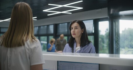 Adult woman stands near reception desk in clinic lobby area and comes to appointment with doctor. Female administrator talks with patient at information counter. Medical staff work in modern hospital.